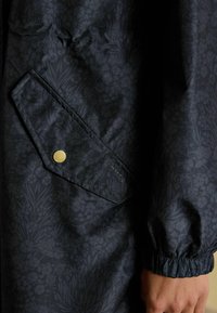 Giacca impermeabile blu navy con motivo floreale, dotata di tasca con bottoni a pressione e polsini elasticizzati per una vestibilità sicura. Materiale strutturato mostrato.