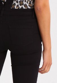 Femme portant un pantalon noir taille haute avec poche arrière et un haut imprimé léopard, bras relâché le long du corps sur un fond uni.