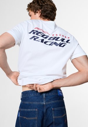 Мъж, носещ бяла тениска Red Bull Racing и сини дънки, показан отзад с ръце, които регулират ризата си.