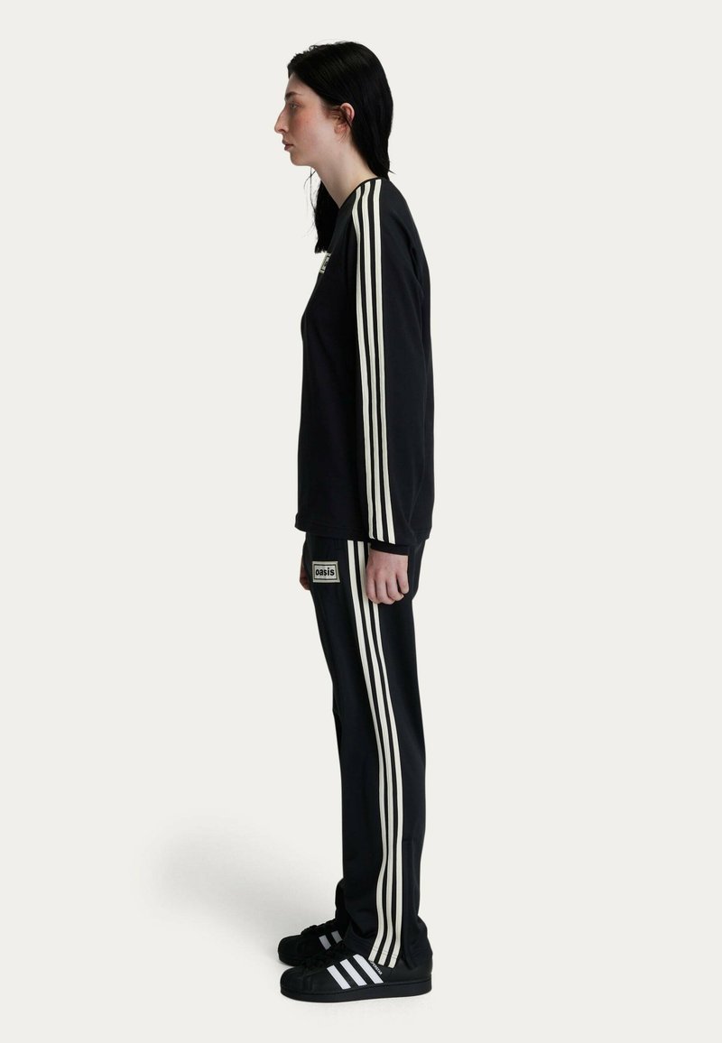 Black tracksuit featuring three white vertical stripes on the sleeves and pants, with a logo patch on the left hip. Worn with black sneakers.