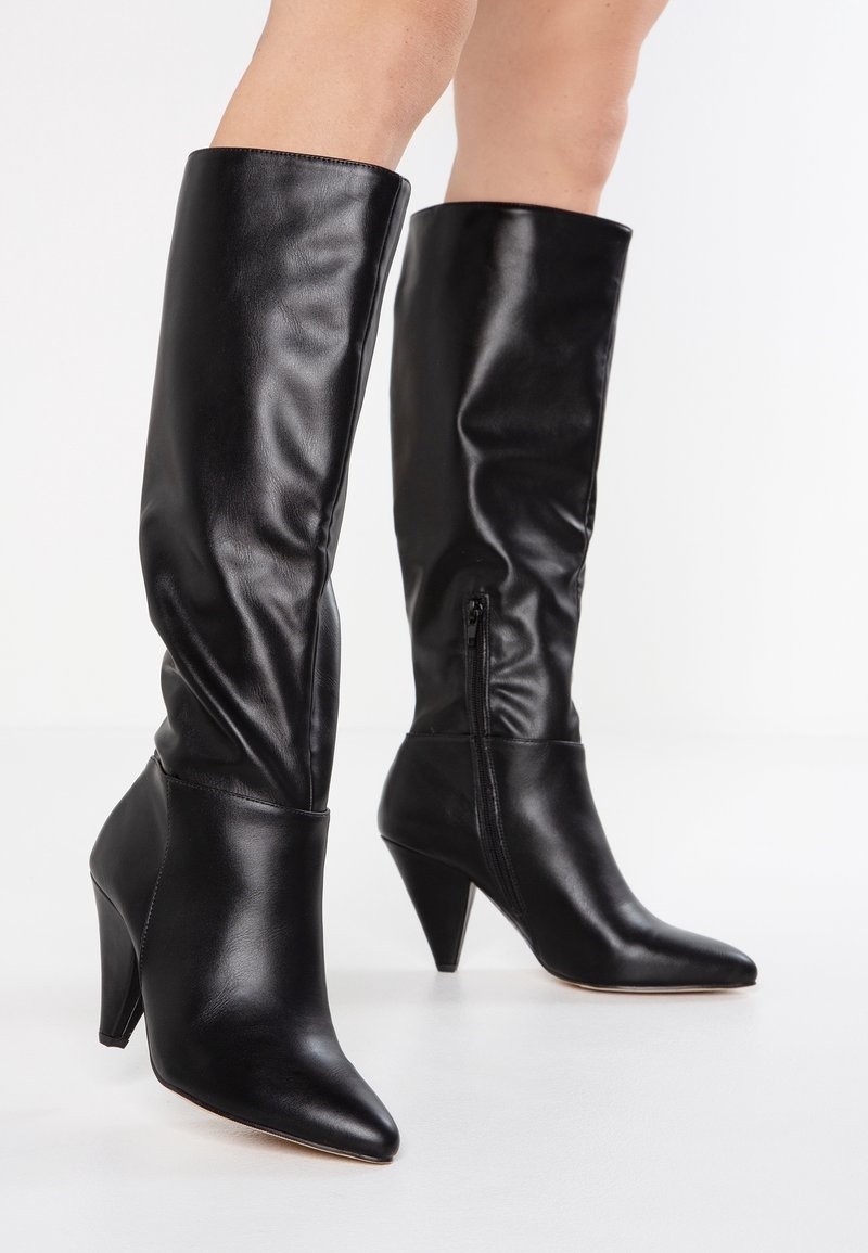 Schwarze kniehohe Lederstiefel mit spitzen Zehen, konischen Absätzen und seitlichen Reißverschlüssen, getragen auf nackten Beinen vor einem weißen Hintergrund.