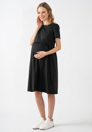 Schwarzes Schwangerschaftskleid mit kurzen Ärmeln, gerafftem Bund und knielangem Saum. Getragen mit weißen Sneakers vor einem neutralen Hintergrund.