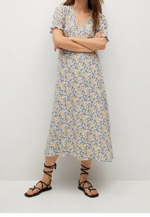 Kobieta w midi kwiecistej sukience z bufiastymi rękawami i czarnych płaskich sandałach na sznurowadła, stojąca z założonymi ramionami na jednolitym tle.