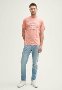 Camiseta de algodón rosa con texto de logo impreso en blanco, combinada con jeans azul claro y zapatillas blancas, de pie contra un fondo claro.