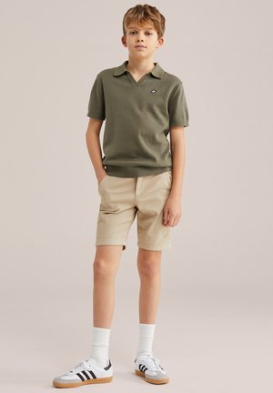 Băiat în picioare, purtând tricou polo verde măsliniu, pantaloni scurți bej, șosete albe și adidași albi cu dungi negre, pe fundal simplu.