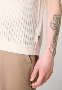Un haut en tricot beige clair avec des côtes verticales, un col rond et une petite étiquette de marque sur le côté, associé à un pantalon marron.