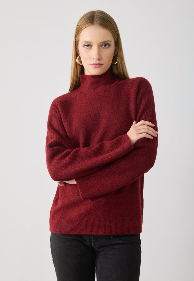 Burgundi turtleneck pulóver puha, textúrált kötésből. Hosszú, lecsúsztatott ujjak és laza fazon jellemzi. Fekete nadrággal viselve.