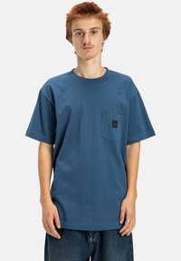 Blue cotton t-shirt with a round neckline and short sleeves, featuring a chest pocket with a black logo label. The fit is relaxed and casual.