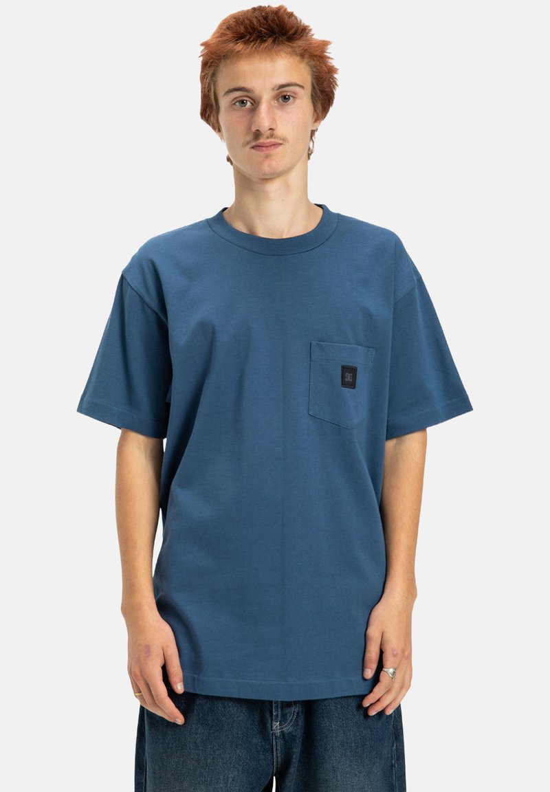 Blue cotton t-shirt with a round neckline and short sleeves, featuring a chest pocket with a black logo label. The fit is relaxed and casual.