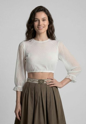 Jonge vrouw met golvend donker haar, draagt een doorschijnende witte cropped blouse en een hoge bruine geplooide rok met decoratieve riem, glimlachend.