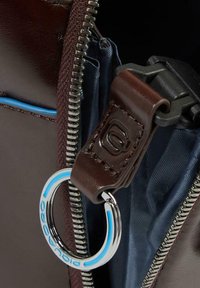 Primo piano di una borsa in pelle marrone parzialmente aperta, che mostra una cerniera metallica, interno in tessuto blu e un portachiavi con il marchio "Piquadro".