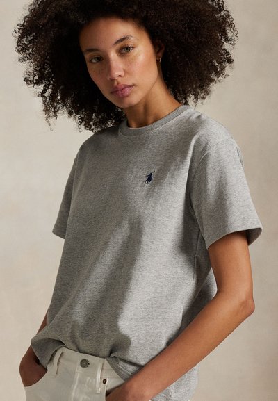 Žena s kučeravými vlasmi nosí sivé tričko s malým tmavým logom na hrudi, zastrčené do bielych nohavíc, s rukami v kapse.