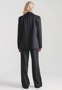 Czarny, dopasowany garnitur z długą marynarką i szerokimi spodniami. Gładka faktura, strukturalny krój i czyste linie. Widok od tyłu.