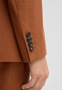 Brauner, strukturierter Blazerärmel mit vier dunklen Knöpfen und kontrastierenden Nahtdetails. Der Stoff wirkt weich und maßgeschneidert.