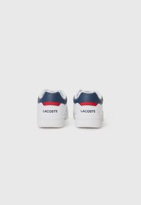 White sneakers with a navy blue and red heel accent, featuring the Lacoste logo in black. Smooth leather exterior with textured rubber sole.