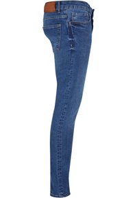 Jean slim en denim bleu vu de côté droit, avec cinq poches, passants pour ceinture et un écusson de marque en cuir marron sur la ceinture.