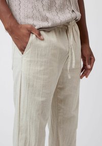 Pantalons en lin beige clair avec une coupe décontractée, présentant une texture verticale visible, une taille avec cordon de serrage et des poches latérales.
