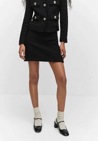 Femme portant un tailleur jupe noir texturé à deux pièces avec des boutons dorés, des chaussettes blanches et des escarpins Mary Jane noirs laqués, se tenant devant un fond blanc.