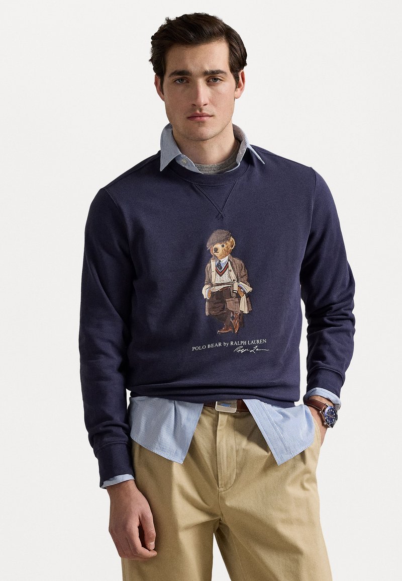 Sweatshirt bleu marine à encolure ronde, comportant un graphique d'un ours en costume. Superposé à une chemise à col bleu clair, associé à un pantalon beige.