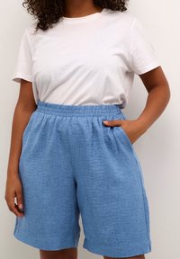 Pantaloncini azzurri con tessuto morbido e testurizzato e cintura elastica. Abbinati a una maglietta bianca aderente e liscia. Mani nelle tasche.