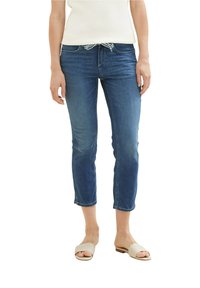 Mujer vistiendo unos jeans cortos azules con cintura ajustable, una blusa sin mangas blanca y sandalias beige sobre un fondo blanco.