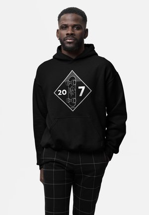 Hombre con sudadera con capucha negra con un gráfico de monopatín y los números 20 y 7, acompañado de pantalones negros con un patrón de líneas de cuadrícula blancas.
