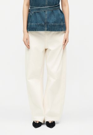 Person, die cremefarbene, weit geschnittene Hosen, schwarze spitze Schuhe und ein ärmelloses Jeansoberteil mit Gürtel in der Taille trägt, vor weißem Hintergrund stehend.