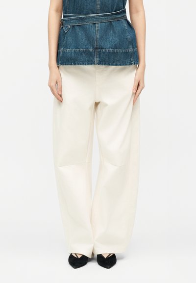 Persona vestida con pantalones anchos de color crema, zapatos negros puntiagudos y una blusa sin mangas de mezclilla con un cinturón alrededor de la cintura, de pie contra un fondo blanco.
