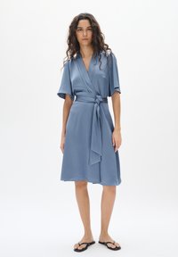Blaues Wickelkleid mit strukturierter Oberfläche, kurzen Ärmeln und einem gebundenen Taillendetail. Kombiniert mit schwarzen Flip-Flops, vor einem weißen Hintergrund.