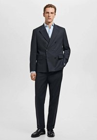 Blazer grigio scuro doppiopetto con bottoni neri, abbinato a pantaloni slim-fit. Camicia azzurra con colletto e scarpe nere. Texture liscia.