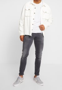 Chaqueta de mezclilla blanca con dos bolsillos en el pecho y botones marrones, llevada sobre una camiseta blanca. Pantalones gris ajustados y zapatillas gris claro.