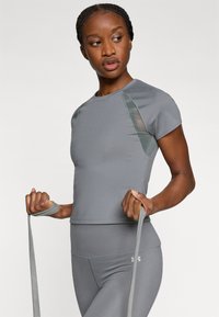 Sivi sportski top kratkih rukava s mrežastim detaljima na ramenima i glatkom teksturom. Model drži trake za otpor u neutralnom stavu.