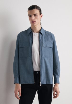 Camicia con zip in tessuto azzurro chiaro, dotata di due tasche sul petto, colletto e maniche lunghe, indossata sopra una maglietta bianca e pantaloni neri.