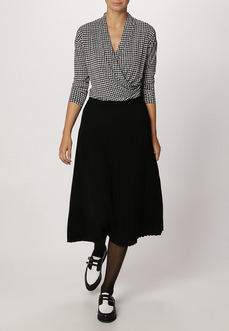 Femme portant un haut cache-cœur à motif pied-de-poule noir et blanc, une jupe midi plissée noire, des collants noirs et des mocassins noir et blanc marchant vers l'avant.