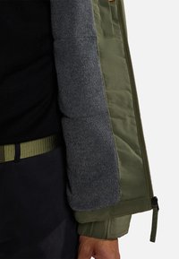 Giacca imbottita con esterno in nylon verde oliva e interno in felpa grigia, con tessuto nero sottostante e una cintura strutturata visibile.