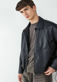 Giacca di pelle nera con collare, chiusura con zip sul davanti e due tasche laterali. Indossata sopra una maglietta grigia e pantaloni grigi con texture.