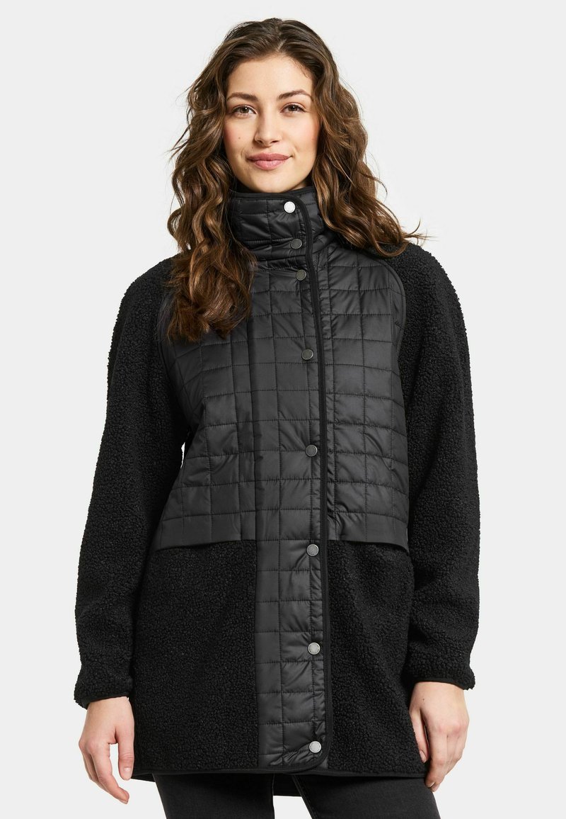 Kvinna som bär en svart vadderad och fleece lång jacka med tryckknappar, stående mot en enkel vit bakgrund.