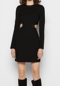 Femme portant une mini-robe noire à manches longues avec des découpes rondes à la taille et des bottes noires montantes contre un fond blanc.