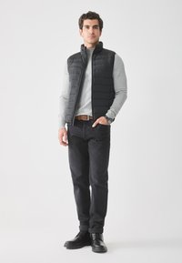 Chaleco acolchado negro sobre una camiseta de manga larga gris, jeans oscuros y botas negras. Los detalles notables incluyen un cuello alto y cierre con cremallera.