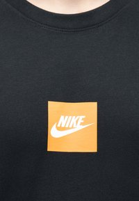 Μαύρο T-shirt με στρογγυλή λαιμόκοψη και ένα πορτοκαλί τετράγωνο στο κέντρο, στο οποίο βρίσκεται το λευκό λογότυπο της Nike και το swoosh στο στήθος.