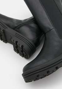 Les bottes noires en cuir haute jusqu'au genou présentent une surface texturée, des panneaux latéraux élastiques et une semelle en caoutchouc robuste avec une bande de roulement à motif pour une meilleure traction.