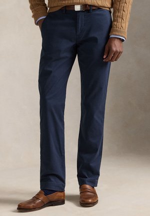 Man met marineblauwe broek, bruine riem, bruine leren loafers en een beige kabelgebreide trui met zichtbare lichtblauwe overhemdsmouwen.