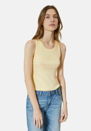 Młoda kobieta z długimi włosami, ubrana w jasny żółty top bez rękawów i niebieskie dżinsy, stoi na tle jednolitego białego tła.