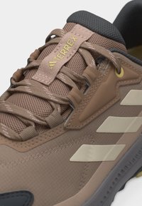 Καφέ αθλητικό παπούτσι Adidas Terrex με μπεζ ρίγες, υφαντό ύφασμα, υφή στη σόλα και κορδόνια με θηλιές σε λευκό φόντο.