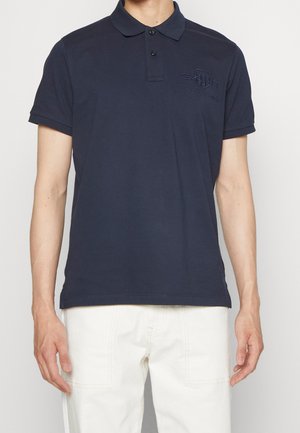 Homme portant un polo bleu marine à manches courtes avec un col à deux boutons et un pantalon blanc, debout devant un fond uni.