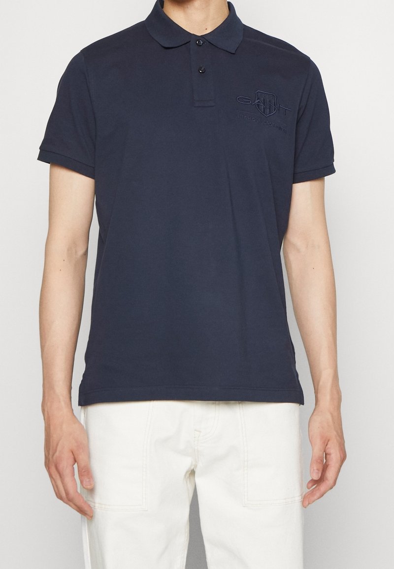 Homme portant un polo bleu marine à manches courtes avec un col à deux boutons et un pantalon blanc, debout devant un fond uni.