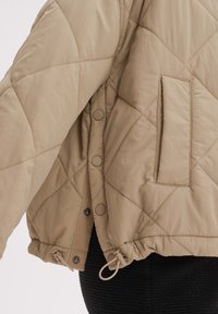 Beżowa pikowana kurtka z zatrzaskami, boczną kieszenią i regulowanym ściągaczem na dole, noszona na teksturowane czarne spodnie.