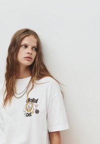 T-shirt en coton blanc avec un design graphique d'un animal de compagnie numérique, orné d'accents colorés et de texte. Manches courtes et coupe décontractée.