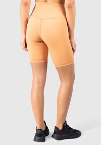 Vysoké broskvové cyklistické šortky vyrobené z elastického materiálu, s hladkým povrchem a bezešvým designem, spárované s černými sportovními botami.