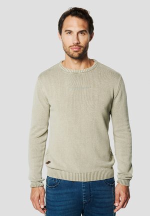 Jersey de punto gris claro con cuello redondo y puños acanalados, con un sutil logo bordado en el pecho. Combinado con jeans azules.